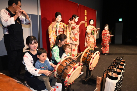 じゅうまりゅうまつりばやしの太鼓を、来場した子どもが体験している様子。隣でおかめさんが踊っている。