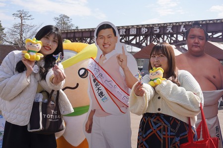 所沢市観光大使のお笑い芸人・春日さん、力士・北勝富士関のパネルの前で、辰巳雄大さんのアクリルキーホルダーを持った女性二人がカメラに笑顔を向ける様子。パネルの後ろにはトコろんが写りこんでいます。